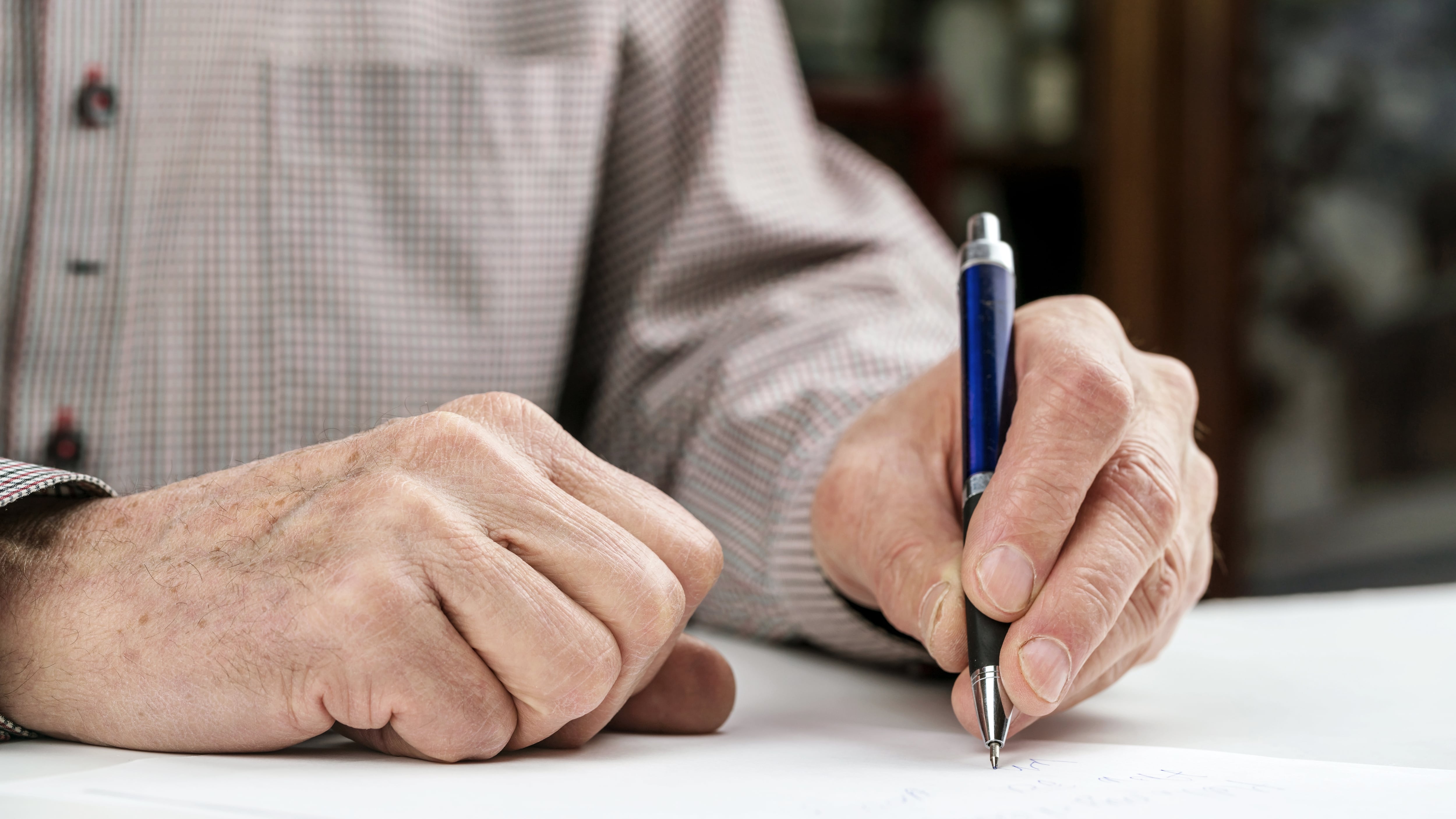senior man in grey shirt tries to write letter with left hand using ball pen at table in light room at home close view